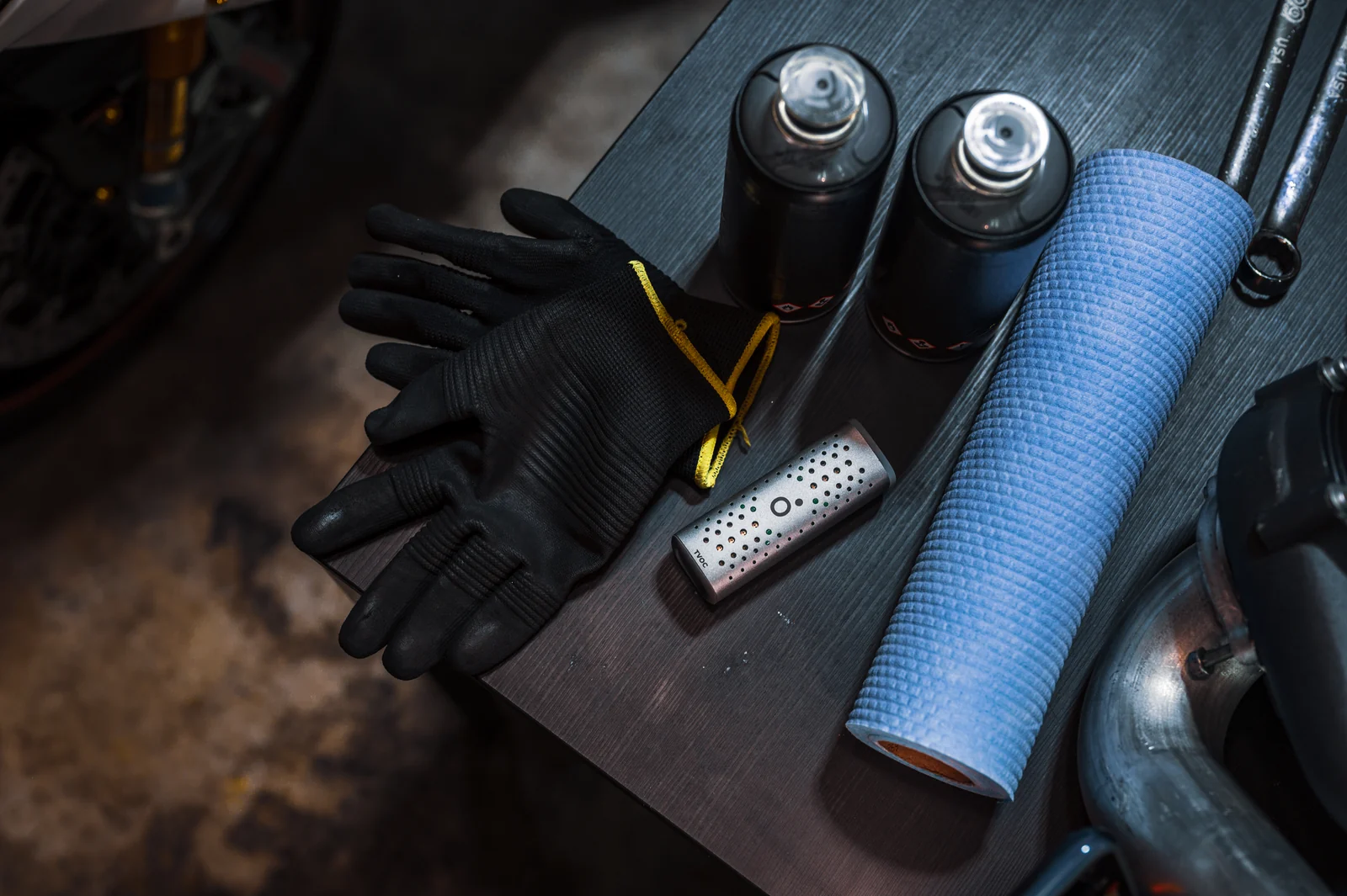 An Airo TVOC Pod lying on a workbench beside gloves, aerosol cans, a blue cloth roll and wrenches.