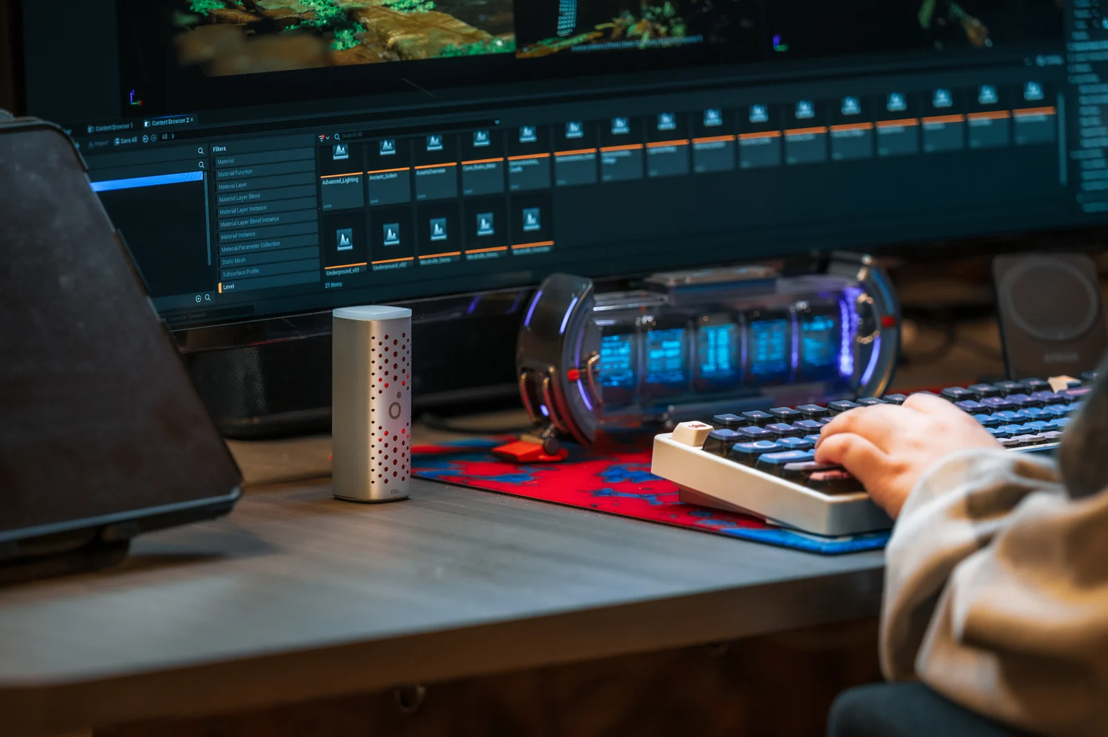 An Airo CO₂ Pod beside a keyboard at a creative workstation, with a person working at a large monitor.